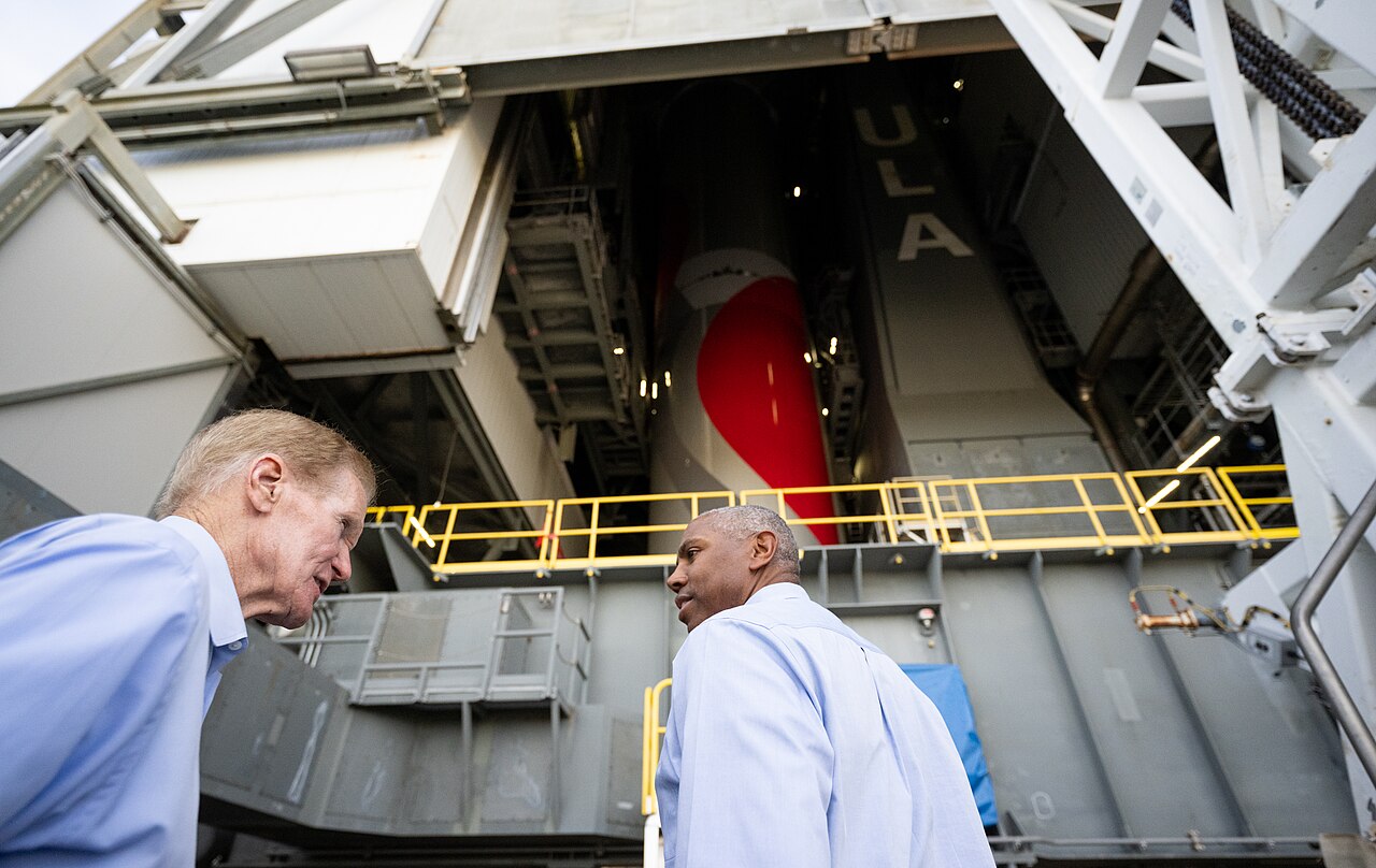 Two men stand in front of a large white rocket inside a tall building.